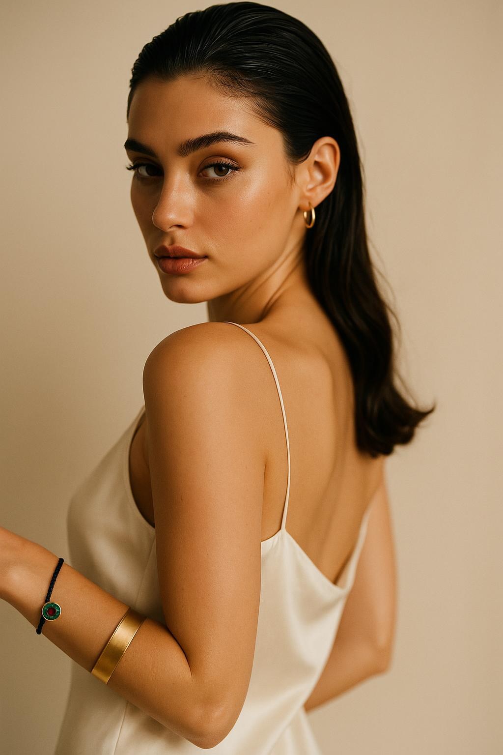 Woman in a pearl silk slip dress looking over her shoulder, wearing the FOUTNI Black Edition braided cord bracelet with a gold evil eye charm